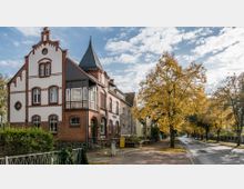 Historisches Wohnhaus aus rotem Ziegel mit detaillierten weißen Fassadenelementen und einem spitzen Turmdach; daneben eine von Bäumen gesäumte Straße mit herbstlich gelbem Laub auf den Bäumen und Gehwegen.