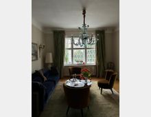 Wohnzimmer mit einem klassischen Stil. Im Mittelpunkt steht ein runder Glastisch mit gelben Sesseln und einer Vase mit roten Blumen, daneben ein dunkelblaues Sofa. Ein Kronleuchter hängt von der Decke, und durch ein großes Fenster mit Sprossen fällt Tageslicht, eingerahmt von grünen Vorhängen.