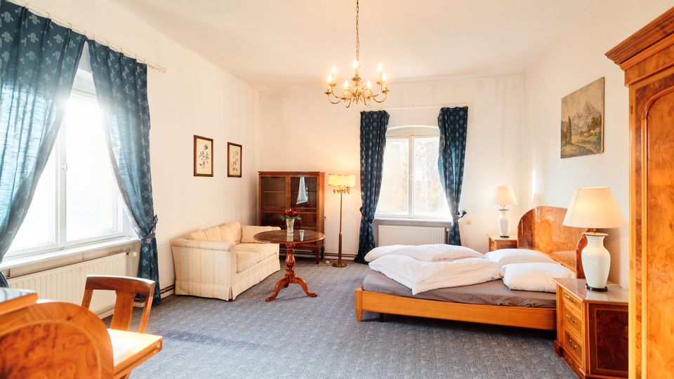 Helles Schlafzimmer mit Doppelbett aus Holz, Nachttischen und Tischlampen sowie einem kleinen Sitzbereich mit Sofa und rundem Beistelltisch. Große Fenster mit dunkelblauen Vorhängen, Kronleuchter an der Decke und klassisches Holzmobiliar wie Schrank und Vitrine prägen den Raum.