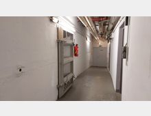 Ein weiß gestrichener unterirdischer Flur mit Betonboden, sichtbaren Rohren an der Decke und einer schweren Metalltür an der linken Seite. Am Ende des Gangs befindet sich eine graue Tür mit einem Notausgangsschild darüber, während an der Wand ein Feuerlöscher montiert ist.