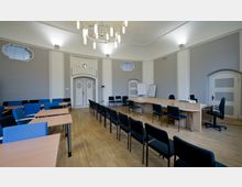 Ein heller Seminarraum mit Holzfußboden, bestuhlt mit blauen und schwarzen Stühlen sowie Tischen. Im Hintergrund befindet sich ein geschwungenes Rednerpult aus Holz, eine doppelflügelige weiße Tür mit Verzierungen und ein moderner Kronleuchter an der Decke.