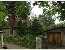 Ein Wohnhaus mit verputzter Fassade und Ziegeldach, teilweise verdeckt durch Bäume und Sträucher, daneben eine Garage mit Holzflügeltüren. Im Vordergrund befindet sich ein niedriger Lattenzaun und ein gepflasterter Gehweg.