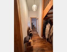 Schmaler Flur in einem Wohnhaus mit Holzboden und weißen Wänden, beleuchtet von einer hängenden Kugellampe. Rechts sind eine Holztreppe und eine Garderobe mit Kleidung, links befinden sich ein Hocker und gestapelte Gegenstände.