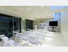 Heller Seminarraum mit weißen Wänden und moderner Einrichtung, darunter weiße Stühle in Reihen angeordnet, ein Regal mit Büchern und ein großer Bildschirm auf einem Rollständer. Ein Stehtisch mit weißem Bezug steht neben einer Glaswand, die in einen angrenzenden Bereich führt.