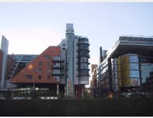 Moderne Bürogebäude mit auffälliger Architektur, darunter ein rotes Backsteingebäude mit schrägem Dach, runden Glastürmen und metallischen Fassadenelementen. Im Vordergrund sind Grünflächen zu sehen, während der Himmel klar ist.