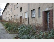 Mehrstöckiges Wohngebäude in einer einfachen Siedlung, mit einer Fassade aus beigem Putz und Eingängen, die von rotbraunen Ziegeln umrahmt sind. Entlang des gepflasterten Weges vor dem Gebäude befinden sich niedrige Büsche und kahle Sträucher.