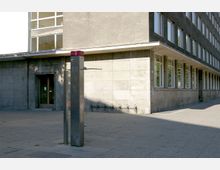 Ecke eines schlichten, grauen Bürogebäudes mit mehreren Fenstern. Im Vordergrund steht eine freistehende, schmale Metallsäule mit einem Telekommunikationslogo, daneben ein Fahrradständer auf dem gepflasterten Platz.