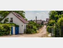 Eine ländliche Gasse mit unbefestigtem Weg, gesäumt von kleinen Häusern mit Gärten und Zäunen. Im Vordergrund ein weißes Haus mit roten Dachziegeln und blauen Türen, im Hintergrund ein Radfahrer und weitere Gebäude.