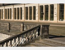 Historische Terrasse mit Balustrade im Vordergrund, dahinter eine Fassade mit hohen Sprossenfenstern und Nischen, die weiße Skulpturen enthalten. Der Gebäudestil weist klassische Elemente und symmetrische Gestaltung auf.