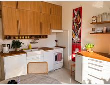 Helle Küche mit weißen Unterschränken, Holzarbeitsplatten und hölzernen Hängeschränken. Eine Spüle, ein kleiner Herd und eine rote abstrakte Malerei an der Wand sind sichtbar, sowie offene Regale mit Küchenutensilien und ein Kräutertopf.