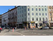 Straßenszene mit mehrstöckigen Wohn- und Geschäftsgebäuden, teils mit Graffiti und Plakaten bedeckt. Im Vordergrund sind Fußwege, parkende Autos, Fahrräder und ein Zebrastreifen zu sehen.