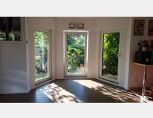 Heller Innenraum mit drei großen bodentiefen Fenstern, die Blick ins Grüne bieten; auf der rechten Seite steht eine Kaffeemaschine auf einem kleinen Tisch, der Boden ist aus Holz.