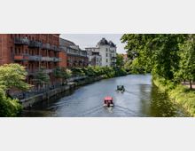 Ein ruhiger Kanal mit zwei kleinen Booten, gesäumt von Bäumen und Gebäuden, darunter ein rotes Backsteinhaus mit Balkonen auf der linken Seite und ein weißes historisches Gebäude im Hintergrund. Entlang des Ufers verläuft ein Gehweg mit Bänken und Geländern.