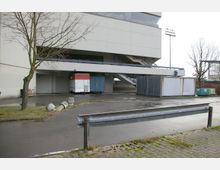 Außenbereich eines modernen Gebäudes mit Betonfassade, einer offenen Treppe und einem überdachten Bereich, in dem Container und ein Verkaufsanhänger abgestellt sind. Im Vordergrund befinden sich ein kahler Baum, Betonpoller, Asphaltflächen und eine Metallleitplanke.