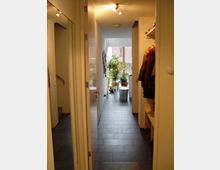 Flur in einem modernen Wohnhaus mit dunklem Fliesenboden, weißen Wänden und Deckenstrahlern. Links führen Treppen nach oben, rechts befindet sich eine Garderobe mit Mänteln, und im Hintergrund steht eine Pflanze vor einem großen Fenster.