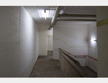 Treppenhaus in einem einfachen Betongebäude mit weißen Wänden, sichtbaren Rohren an der Decke und fluoreszierender Beleuchtung. Eine Treppe mit rotem Handlauf führt nach unten; der Boden besteht aus grauen Fliesen.
