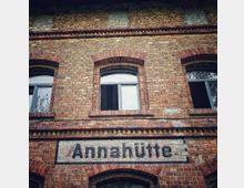 Fassade eines historischen Gebäudes aus rotem Backstein mit rundbogigen Fenstern und der gut lesbaren Aufschrift „Annahütte“. Die Architektur zeigt Zierelemente und gemauerte Details typisch für Industriebauten des 19. Jahrhunderts.