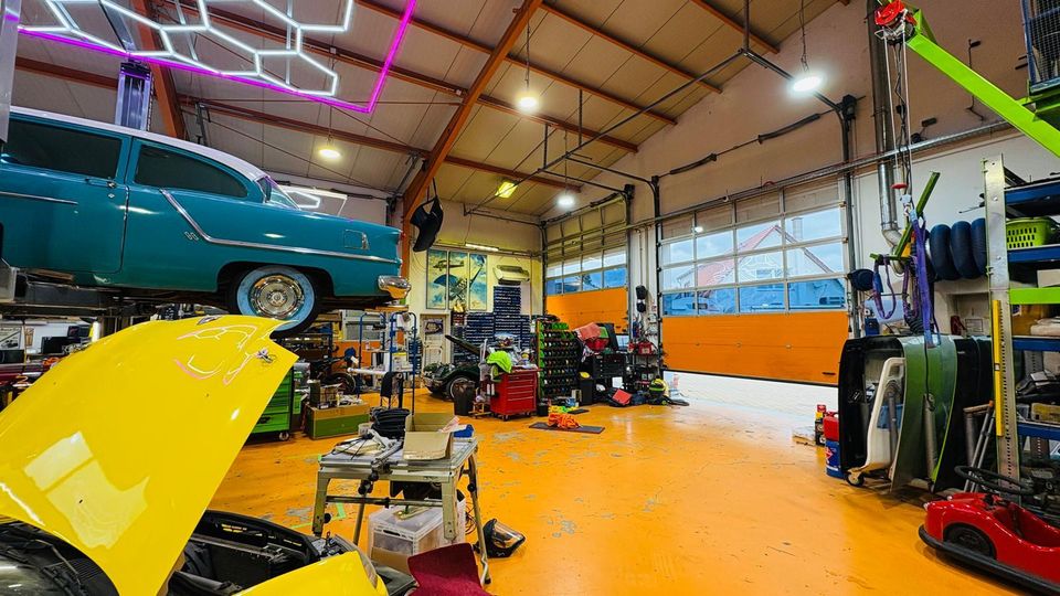 Innenraum einer Autowerkstatt mit orangefarbenem Boden, hohen Deckenbalken und großen Rolltoren mit Fenstern. Zu sehen sind ein Oldtimer auf einer Hebebühne, ein gelbes Auto mit geöffneter Motorhaube sowie zahlreiche Werkzeuge, Regale und Reifen; an der Decke hängen geometrische Neonleuchten.