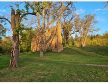 Backsteinruine einer alten Kirche oder Kapelle auf einer grünen Wiese, umgeben von großen, teils blattlosen Bäumen unter einem klaren blauen Himmel. Im Hintergrund dichter Wald und eine leicht hügelige Landschaft.