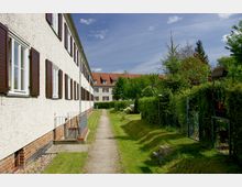 Reihenhaussiedlung mit weiß verputzten Fassaden, braunen Fensterläden und Satteldächern mit roten Ziegeln; ein schmaler Weg führt zwischen dem Gebäude und einer Hecke, die angrenzende Gärten abgrenzt. Im Hintergrund sind weitere ähnliche Gebäude sowie Bäume und Büsche zu sehen.