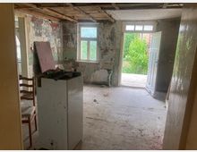 Verlassener Raum mit abgeblätterter Farbe an den Wänden, freiliegendem Holz an der Decke und einer offenen Tür, die nach draußen auf eine kleine Terrasse mit Blick auf üppige Vegetation führt. Im Vordergrund stehen ein Stuhl und ein kleiner weißer Schrank, der teils mit Gegenständen beladen ist.