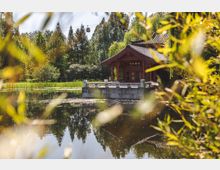 Traditioneller chinesischer Pavillon mit geschwungenem Dach, umgeben von einem ruhigen Teich und dichter Vegetation. Im Hintergrund sind Bäume und eine Seilbahn mit Gondeln sichtbar.