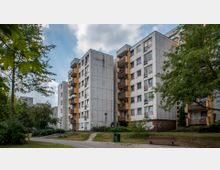 Mehrstöckige Wohngebäude aus Beton mit gelben Balkonen, umgeben von Bäumen und Grünflächen. Im Vordergrund verläuft ein schmaler Gehweg, daneben stehen eine Parkbank und ein Mülleimer.