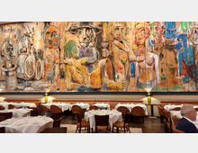 Ein Restaurant mit gedeckten Tischen und gepolsterten Stühlen vor einer großen, farbenfrohen Wandmalerei, die abstrakte Figuren und Tiere zeigt. Die Atmosphäre wird durch dezente Beleuchtung und eine moderne Einrichtung geprägt.