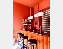 Small bar interior with red-orange walls, a wooden counter lined with black stools, and white globe pendant lights hanging overhead. Shelves behind the bar display bottles and glassware, with a geometric white partition wall on the right.