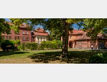Innenhof mit Rasenfläche und großen, schattenspendenden Bäumen, umgeben von Backstein- und Putzgebäuden mit roten Ziegeldächern. Im Hintergrund sind Fensterreihen, ein Torbogen und Hecken entlang eines Weges zu sehen.
