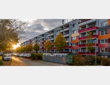 Mehrstöckiges Wohngebäude mit bunten Fassaden in Rot-, Gelb- und Orangetönen, davor parkende Autos entlang einer von Bäumen gesäumten Straße. Im Vordergrund befinden sich Müllcontainer und ein Straßenschild mit der Aufschrift "Fabrikstraße".