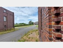 Backsteinfassaden zweier Gebäude an einer asphaltierten Straße, die durch eine Grünfläche mit Bäumen und Sträuchern führt; im Hintergrund ist ein weiteres großes Gebäude erkennbar. Der Himmel ist bewölkt mit vereinzelten blauen Stellen.