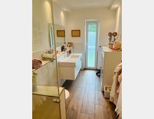 Modernes Badezimmer mit weißer Einrichtung, Holzfußboden und einer schmalen Glastür, die nach außen führt. Links eine Glasduschkabine, in der Mitte ein Wandwaschbecken mit Spiegel, daneben Ablageflächen mit Pflegeprodukten.