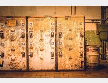 Rostige, industrielle Schaltschränke mit abgenutzter Oberfläche und verrosteten Griffen, in einer Werkstattumgebung mit abgeplatztem Putz an den Wänden und einem Metallfass auf der rechten Seite. Ein Warnschild mit der Aufschrift „Nicht schalten! Lebensgefahr!“ ist sichtbar.