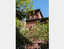 Backsteingebäude mit Relief eines Pferdekopfes an der Fassade, umgeben von grünen Bäumen und blühenden Sträuchern unter klarem blauem Himmel.