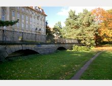 Historisches Gebäude mit einer Fassade im klassizistischen Stil, gelegen neben einer gepflasterten Brücke mit schmiedeeisernem Geländer. Im Vordergrund sind eine weitläufige Rasenfläche und ein schmaler Fußweg sichtbar, umgeben von Bäumen, teils in herbstlicher Färbung.