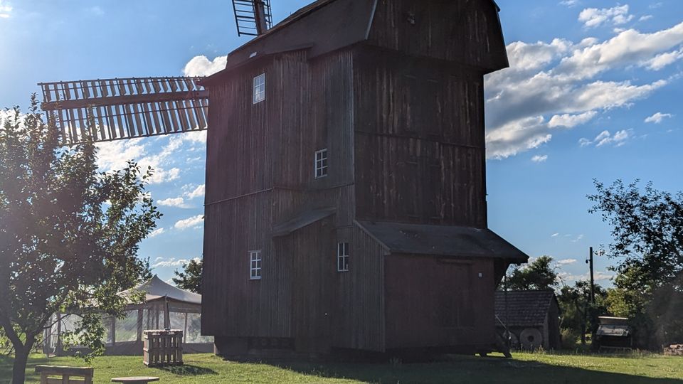 Hohe, dunkle Holzwindmühle mit großen Flügeln auf einer offenen Wiese, aufgenommen vor blauem Himmel mit Wolken. Im Vordergrund stehen Gras, ein kleiner Baum und einfache Sitzgelegenheiten; im Hintergrund sind weitere kleine Gebäude und Zäune zu sehen.