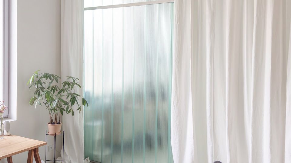 Heller, minimalistisch eingerichteter Innenraum mit großem Fenster bzw. milchiger Glasfront und bodenlangen weißen Vorhängen. Links stehen ein Holztisch und eine Topfpflanze, im Vordergrund sind zwei Stühle auf einem hellen Boden zu sehen.