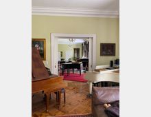 Wohnraum mit klassischem Interieur, dominierend sind zwei Klaviere (ein weißes und ein dunkelbraunes) sowie ein Flügel auf einem roten Teppich. Der Raum hat helle Wände, Holzfußboden, einen Kronleuchter und mehrere gerahmte Gemälde sowie Möbel im Stil der klassischen Einrichtung.