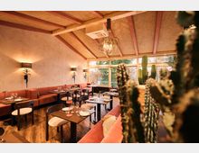 A warmly lit restaurant interior featuring pink cushioned banquette seating along the walls, small wooden tables set with plates and glasses, and a woven ceiling with exposed beams. Large windows allow natural light and provide views of trees, while potted cacti and plants add a tropical decor element.