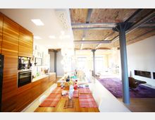 Offener Wohnraum im Loft-Stil mit sichtbaren Backstein-Decken und Metallträgern. Der Raum beinhaltet eine moderne Küche mit Holzfronten, einen Esstisch mit rot karierten Tischsets und einen Wohnbereich mit einem lila Teppich und großen Fenstern, die viel Licht hereinlassen.