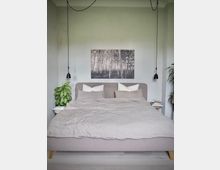 Minimalistisch eingerichtetes Schlafzimmer mit einem grauen Polsterbett, zwei Hängelampen, einem monochromen Bild eines Waldes an der Wand und Zimmerpflanzen auf beiden Seiten des Bettes. Der Raum hat helle Wände und einen Holzboden.