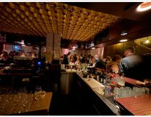 Eine belebte Bar mit einer langen Theke, die mit Gläsern, Flaschen und Barutensilien bestückt ist. Im Hintergrund sitzen und stehen Gäste in einem warm beleuchteten Raum mit einer auffälligen, goldfarbenen Wabenstrukturdecke.