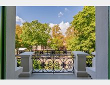 Blick von einem Balkon mit dekorativem schwarzem schmiedeeisernem Geländer und weißen Pfeilern auf eine von großen Laubbäumen gesäumte Wohnstraße. Im Hintergrund sind Wohnhäuser zwischen den Baumkronen unter blauem Himmel zu sehen.