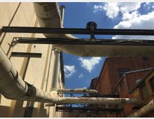 Industrieanlage mit großen, rostigen Rohrleitungen, die an einer beige verputzten und einer roten Backsteinfassade verlaufen. Der blaue Himmel mit vereinzelten Wolken bildet den Hintergrund.