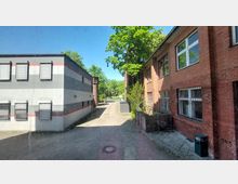 Ein gepflasterter Innenhof, flankiert von einem modernen grauen Gebäude mit horizontalen roten und grauen Streifen auf der linken Seite und einem roten Ziegelgebäude mit vergitterten Fenstern auf der rechten Seite. Im Hintergrund sind Bäume und blauer Himmel sichtbar, die eine grüne Umgebung andeuten.