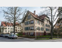 Mehrfamilienhäuser im Stil der frühen 20. Jahrhunderts mit roten Satteldächern, cremefarbenen Fassaden und grünen Fensterläden auf einer Straßenecke; umliegende Bäume und parkende Autos ergänzen die ruhige Wohnumgebung. Ein Straßenschild markiert die Kreuzung von zwei Seitenstraßen.