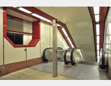 Unterirdischer U-Bahn-Bereich mit Rolltreppen, weißen Wandflächen und roten Akzenten. Beleuchtung erfolgt durch rechteckige Deckenlampen, linke Wand mit einem großen, rot umrahmten Fensterdetail.