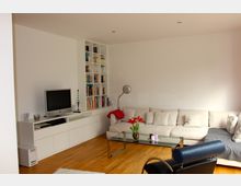 Helles Wohnzimmer mit Holzboden, einer weißen L-förmigen Couch, einem Glas-Couchtisch mit roten Blumen darauf und einem modernen schwarzen Sessel. Im Hintergrund eine weiße Schrankwand mit integriertem Bücherregal, einem Fernseher und weiteren Dekorationselementen.