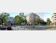 Straßenszene in einer Stadt mit einem fünfstöckigen Eckgebäude im Zentrum, das Balkone und ein Geschäft im Erdgeschoss hat. Bäume und geparkte Autos säumen die Straße, während ein kleiner Kioskbereich links im Bild sichtbar ist.